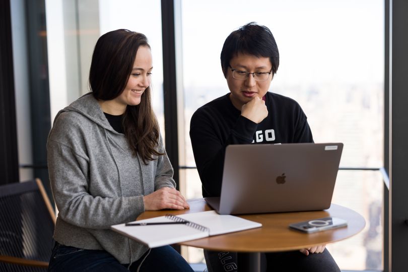 Two people at a table next to a large window. One has a notebook in front of them, the other a laptop. They are both looking at the laptop.